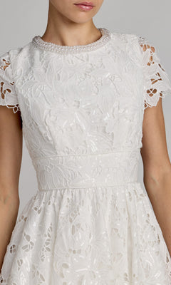 A woman wearing the Mac Duggal 56324 midi party dress is shown from the waist up. The white lace dress features short sleeves, a floral pattern, pearl trim at the neckline, and a fit-and-flare silhouette that flatters her figure.