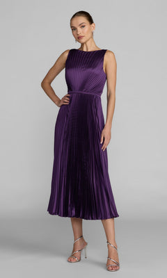 A woman wearing the Mac Duggal 56326 midi party dress in purple, featuring a sleeveless, pleated charmeuse bodice and an elegant A-line skirt, paired with silver strappy heels and standing against a plain light gray background.