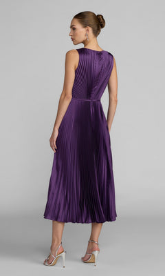A woman stands with her back to the camera against a plain gray background, wearing the Midi Party Dress: Mac Duggal 56326—a sleeveless, pleated charmeuse dress featuring an A-line skirt in a rich purple hue—paired with silver heels.