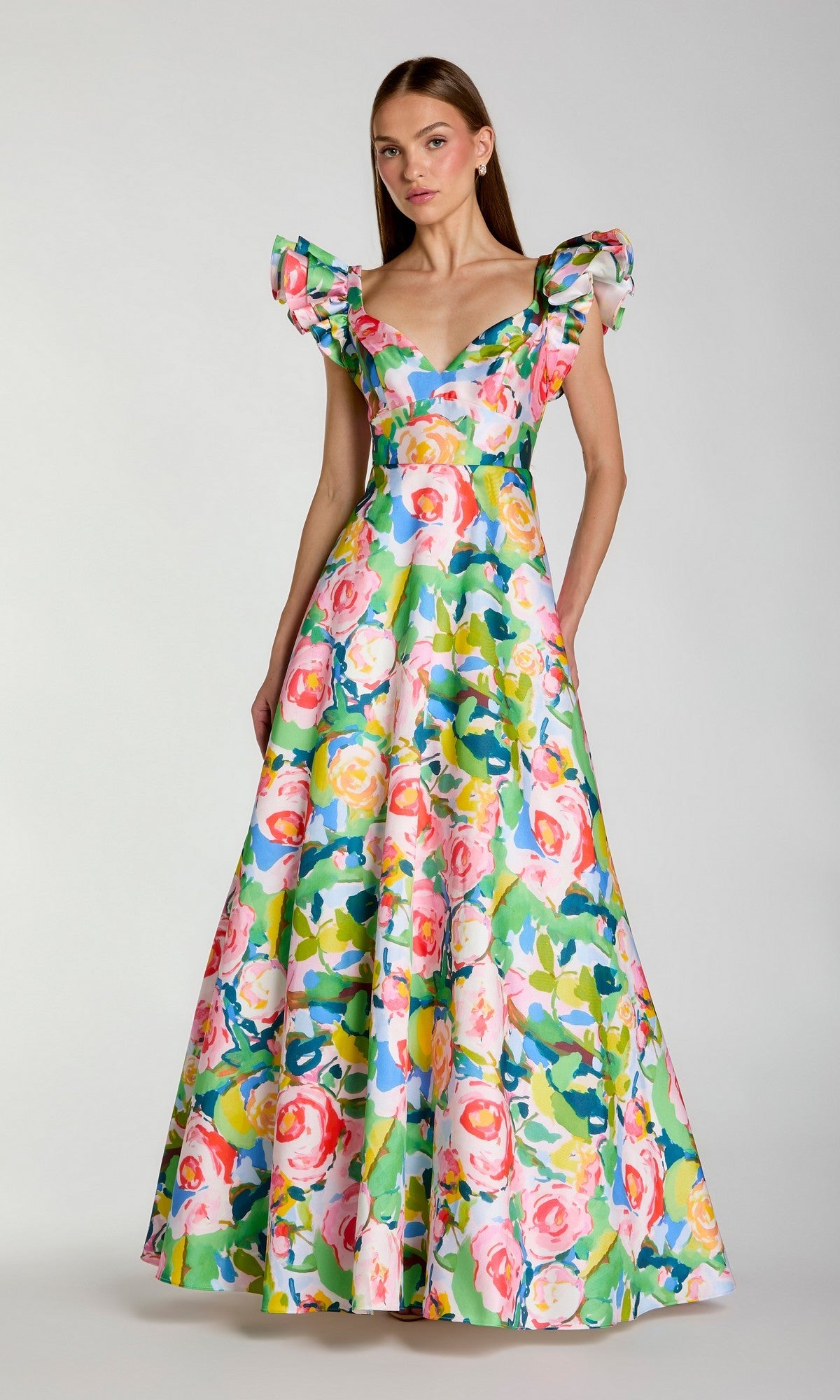 A woman wears the Mac Duggal 56341 Long Formal Dress, a floor-length ballgown crafted from floral Mikado fabric in a vibrant blue multi colorway. The gown features ruffled sleeves and showcases bright hues on a crisp white background.