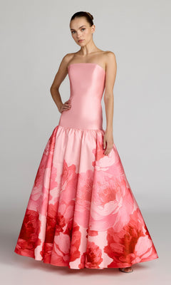 A woman stands in the Mac Duggal 56343 long formal dress, featuring a strapless fitted bodice and a flared skirt with blush multi hues and a red floral pattern near the hem, echoing the brand’s signature elegance.