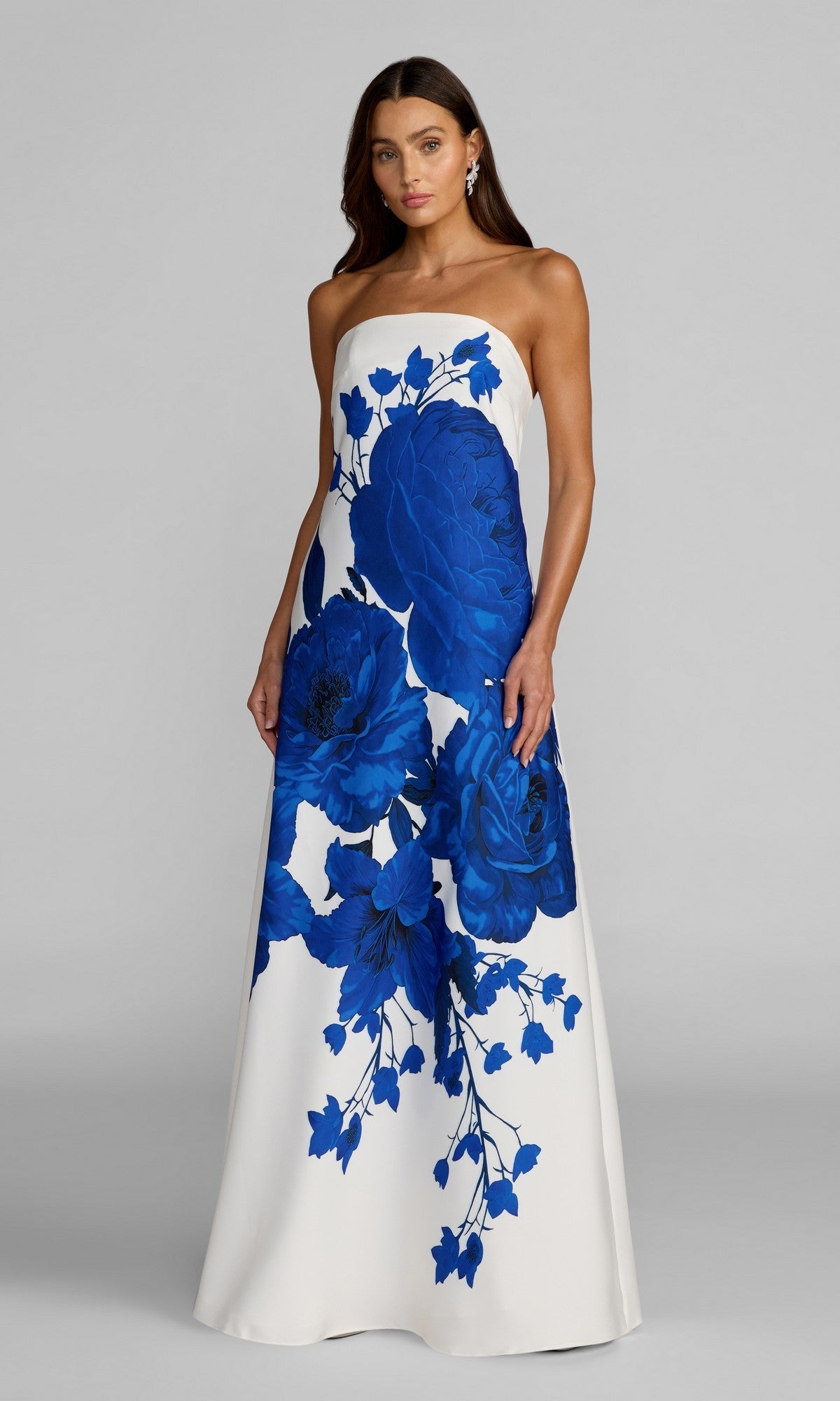 A woman wearing the Mac Duggal 56344 long formal dress in White Multi, featuring a strapless design with a bold blue floral print, stands against a plain gray background.