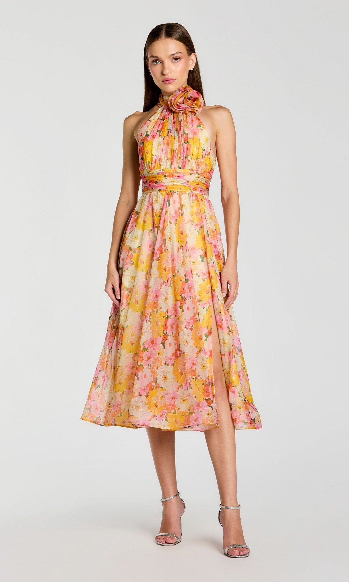 A woman wearing the Mac Duggal 56348 yellow multi sleeveless floral-printed chiffon midi party dress with a halter neck and bow detail, paired with silver high-heeled sandals, stands against a plain light background.