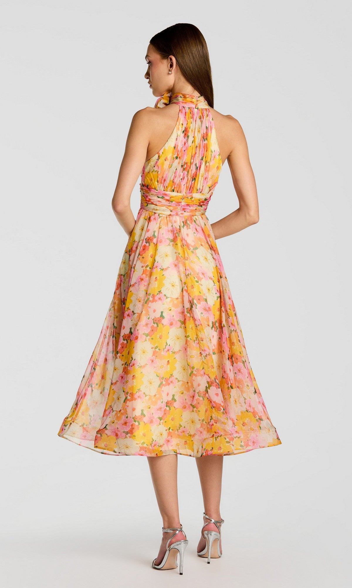 A woman stands with her back to the camera, wearing the Mac Duggal 56348 sleeveless floral-printed chiffon midi party dress with an open back, paired with silver high-heeled sandals against a plain background.