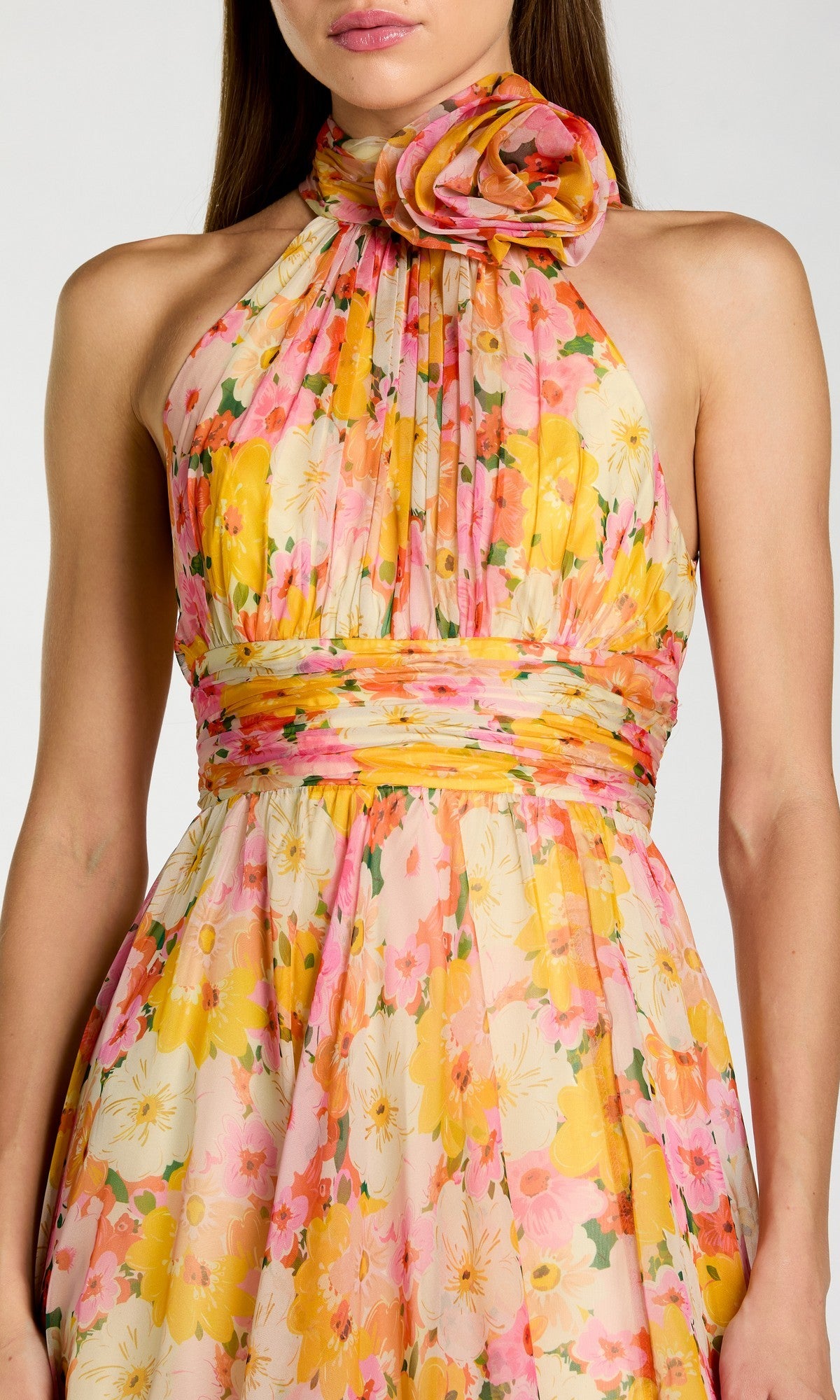 A woman wearing the Midi Party Dress: Mac Duggal 56348, a sleeveless halter-neck dress with yellow, pink, and orange floral prints, featuring a large fabric flower at the neckline and a cinched waist.