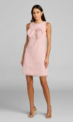 A woman wears the Mac Duggal 56370 Short Boucle Shift Dress in blush—a sleeveless dress featuring pearl embellishments, a statement bow on the chest, and pearl-accented hem—standing against a plain gray background.