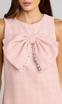 A woman wearing the Mac Duggal 56370 Short Boucle Shift Dress, a sleeveless pink textured dress featuring a large bow with pearl accents at the front.