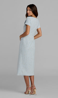 A woman with her back turned, looking over her shoulder, models the Mac Duggal 56374 Tea-Length Party Dress in light blue—a textured, short-sleeved style that falls to the knee—paired with beige high-heeled sandals against a plain background.