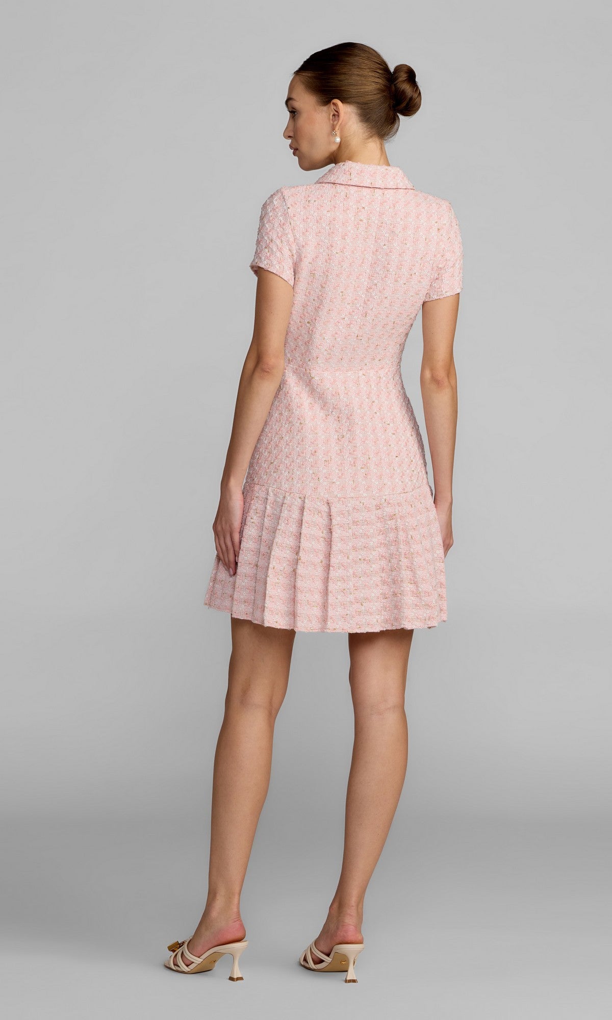 A woman stands with her back to the camera, wearing the Mac Duggal 56379 Short Party Dress—a light pink, short-sleeve A-line dress with a pleated skirt—paired with heeled sandals, set against a plain gray background.