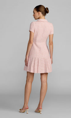 A woman stands with her back to the camera, wearing the Mac Duggal 56379 Short Party Dress—a light pink, short-sleeve A-line dress with a pleated skirt—paired with heeled sandals, set against a plain gray background.