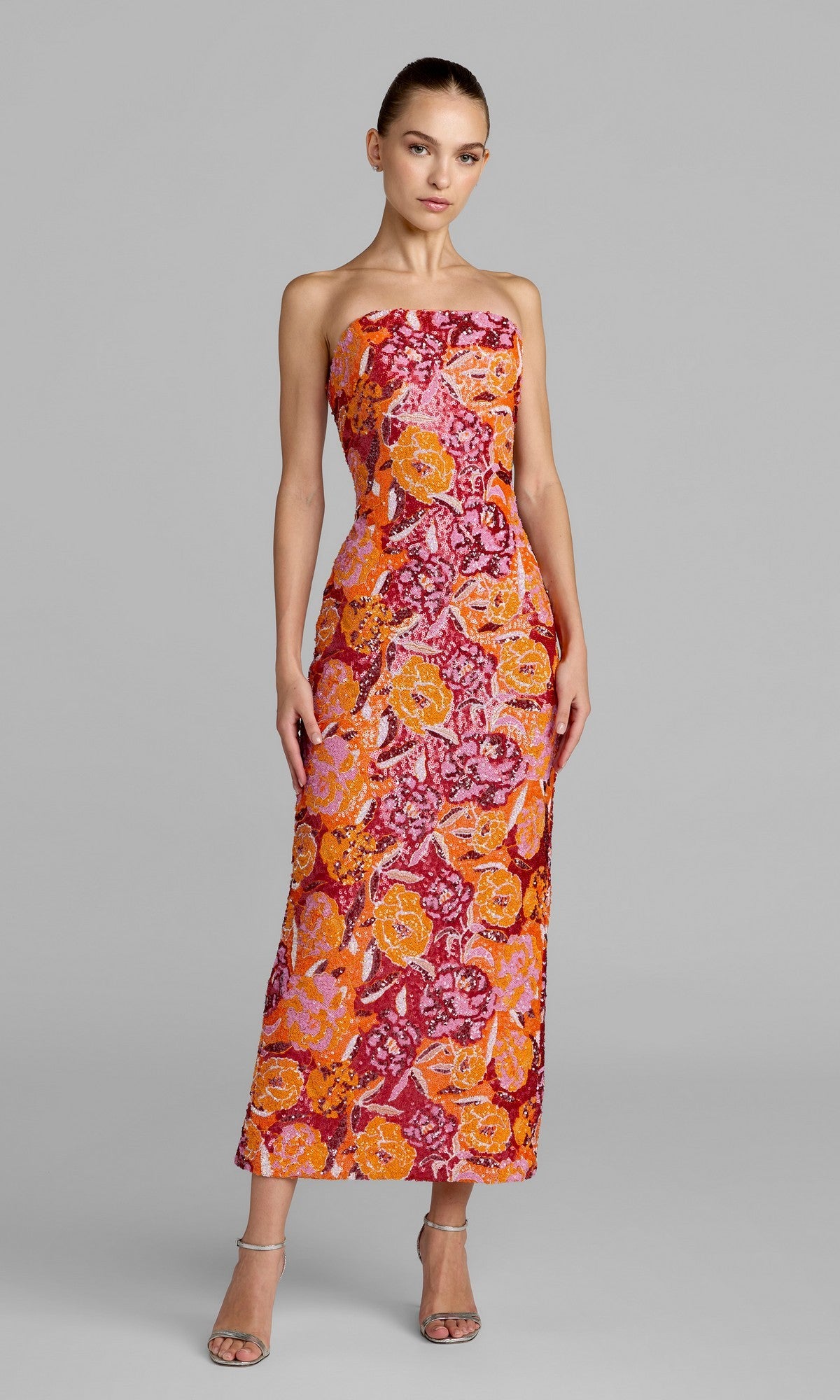 A woman wearing the Mac Duggal 56385 Tea-Length Party Dress in Tangerine Multi, featuring a strapless design with a vibrant orange, pink, and red floral pattern; she stands in strappy heels against a plain gray background.