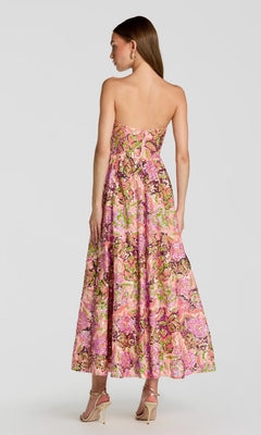 A woman stands facing away from the camera against a plain light background, wearing the Tea-Length Party Dress: Mac Duggal 56386—a strapless, pink floral sequin party dress paired with high heels.