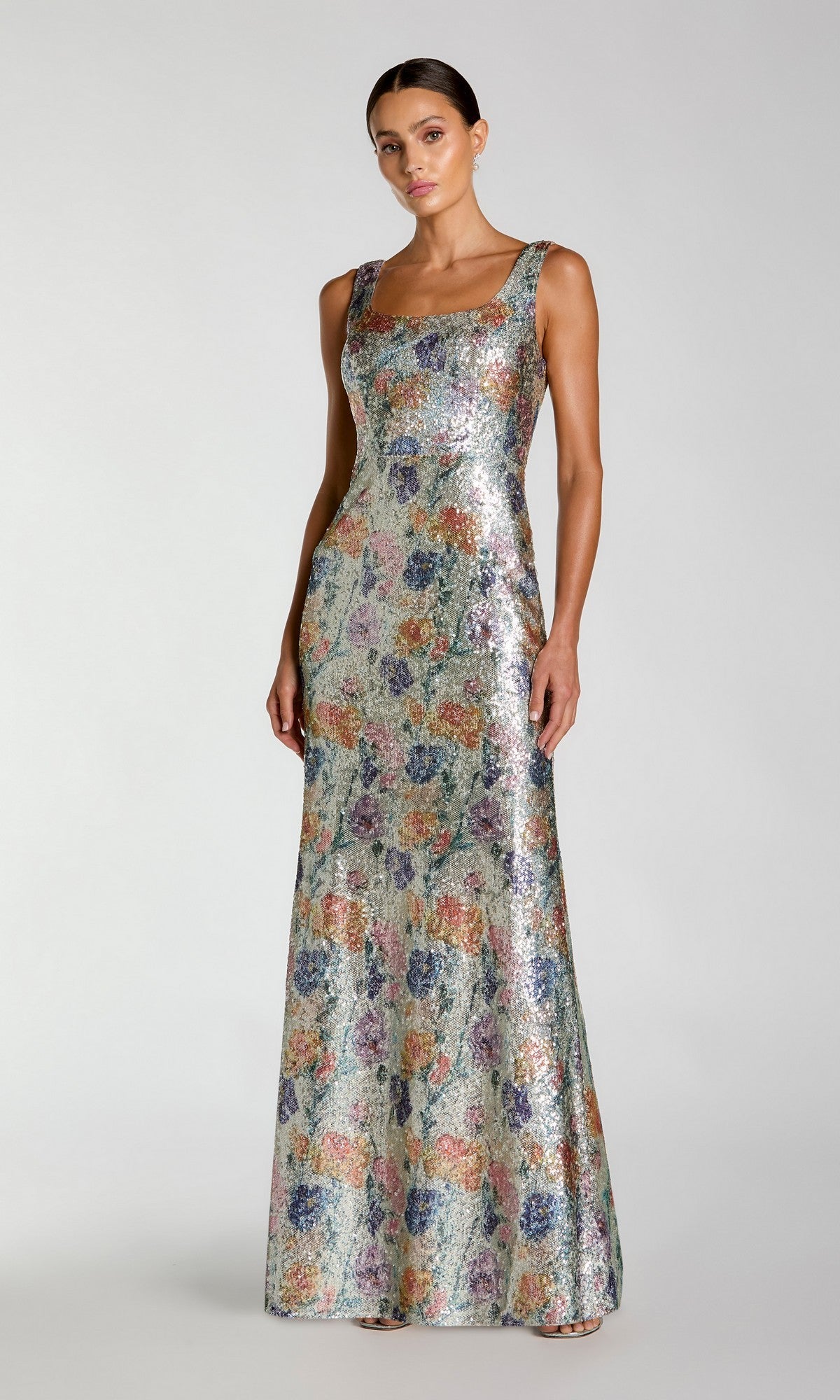 A woman wears the Mac Duggal 56388 Long Formal Dress in Silver Multi—a sleeveless, floor-length fitted gown adorned with colorful floral sequin embellishments—while standing against a plain light gray background.