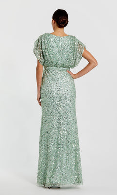 A woman faces away, wearing the Mac Duggal 5640 Short Sleeve Long Sequin Formal Dress—a floor-length, light green gown with sequins, short sleeves, and a belted waist.