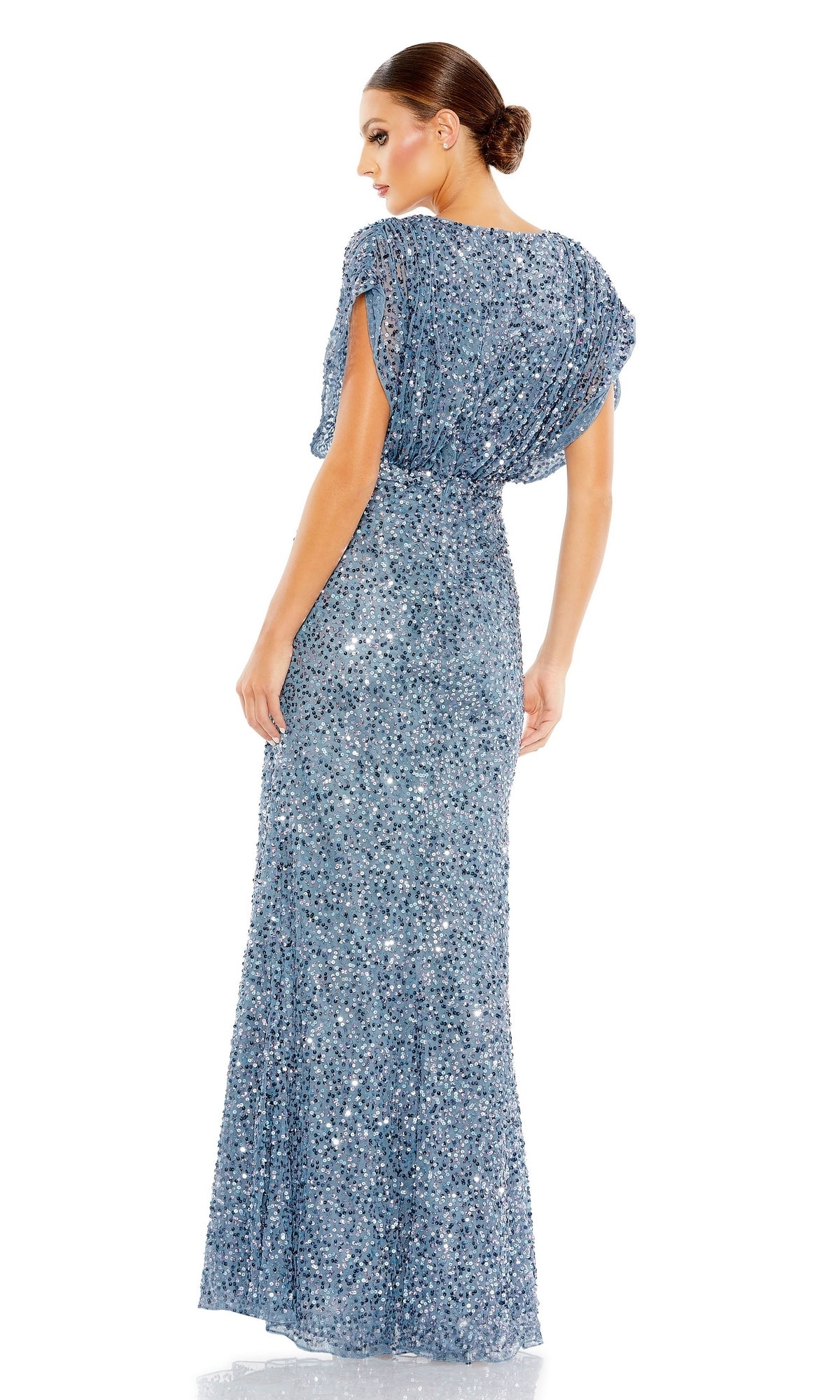 A woman in a Mac Duggal 5640 Short Sleeve Long Sequin Formal Dress stands facing away, glancing over her shoulder, with her hair styled in a low bun.