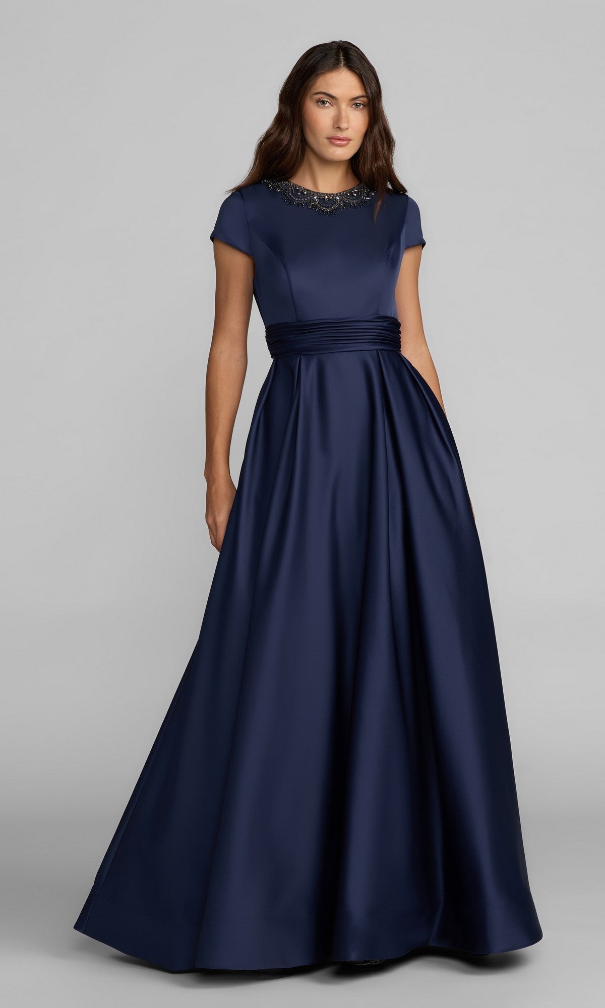 A woman stands against a plain light gray background, wearing the Mac Duggal 56401 long formal dress in Midnight—a navy blue satin gown featuring short sleeves, a beaded neckline, and a fitted waist.