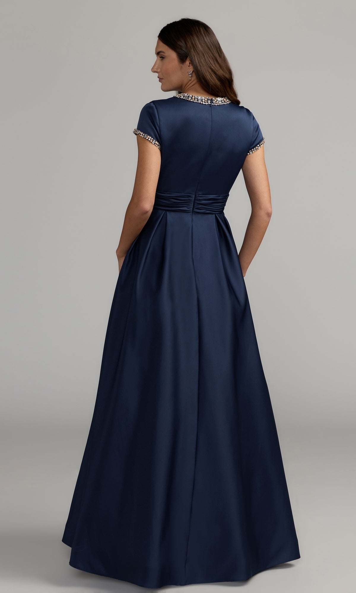 A woman stands facing away against a plain background, wearing the Long Formal Dress: Mac Duggal 56402—a floor-length, short-sleeve navy gown with jeweled trim that evokes timeless elegance.
