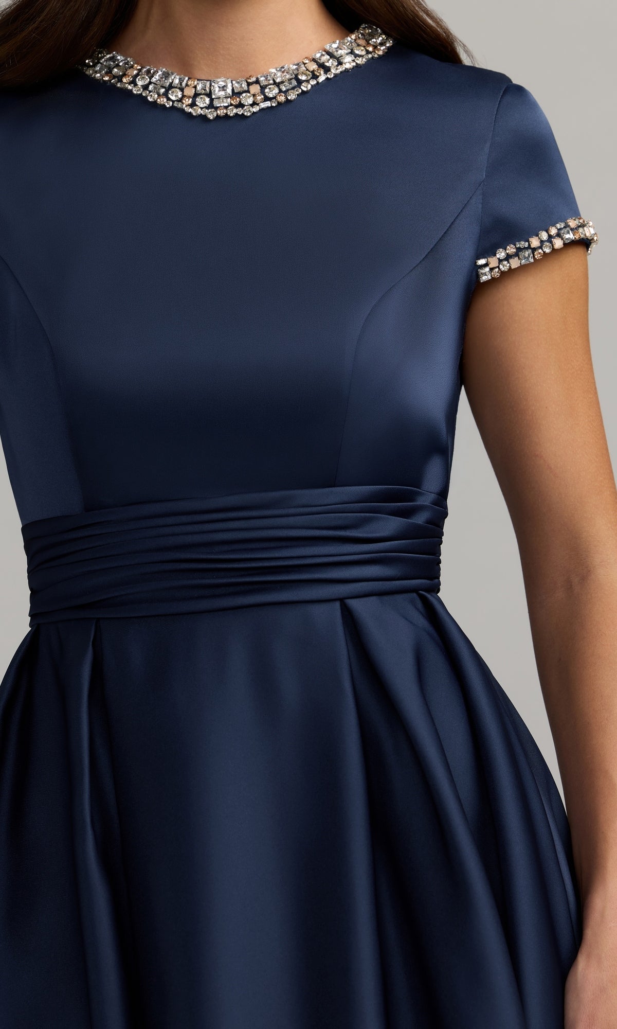 A woman is shown from the waist up wearing the Mac Duggal 56402 long formal dress, featuring a navy blue satin fabric with a jeweled neckline and sleeves, a fitted bodice, ruched waist, and a flared skirt in the style of a classic Mac Duggal satin ball gown.