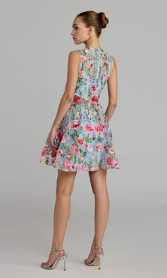 A woman stands in profile against a plain gray background, wearing the Mac Duggal 56409 short party dress—a floral lace mini dress with a fit-and-flare silhouette and a delicate lace back—paired with silver high-heeled sandals.