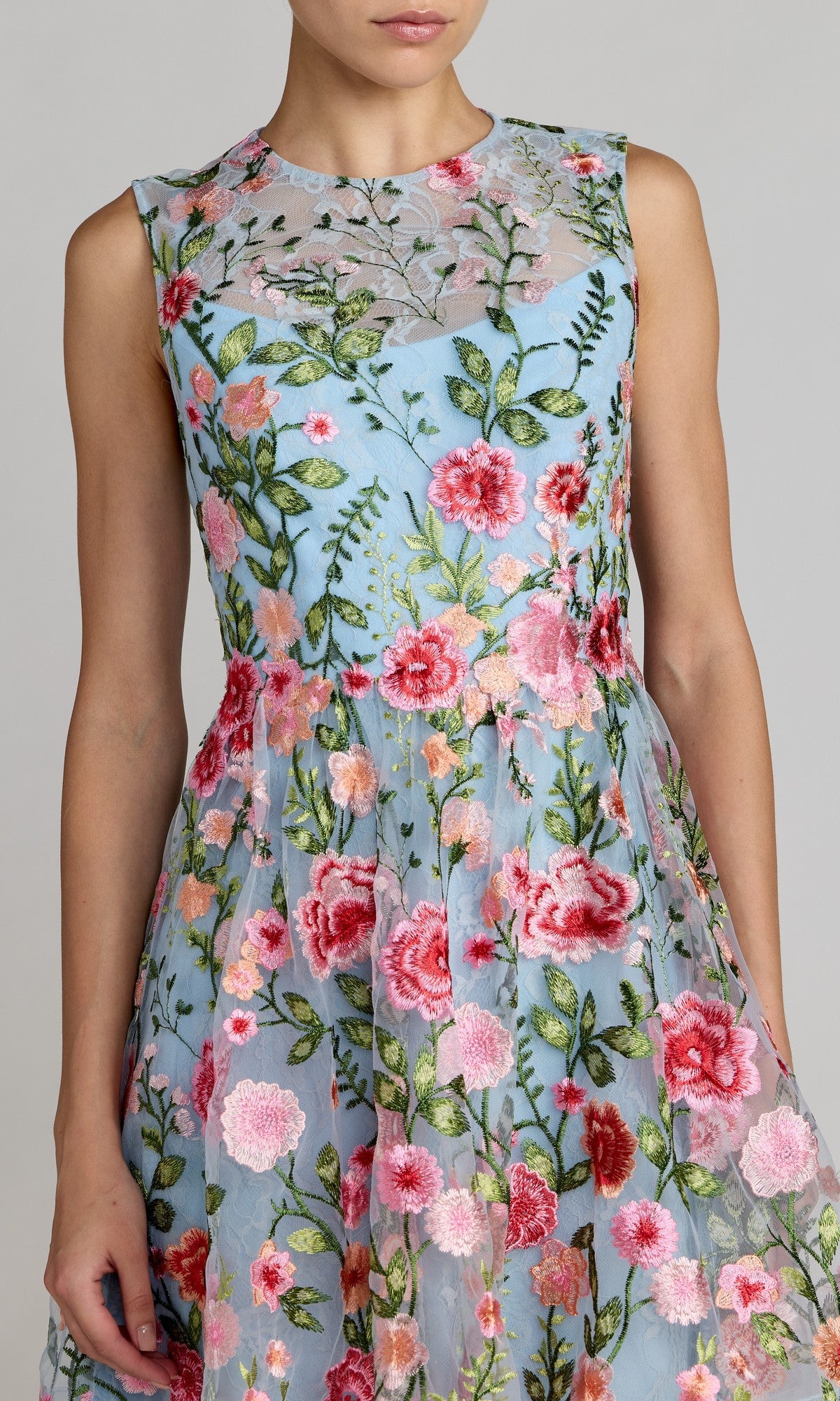 A woman wearing the Mac Duggal 56409 short party dress, featuring a sleeveless light blue floral lace design with pink, red, and green embroidery and a semi-sheer high neckline.