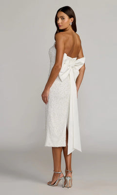 Wearing the Mac Duggal 56416 Midi Party Dress—an elegant, strapless white ensemble featuring a prominent back bow and subtle pearl sequin embellishments—a woman stands in heels, facing away while glancing over her shoulder, epitomizing sophisticated style.