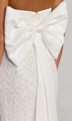 The Midi Party Dress: Mac Duggal 56416 is a white dress with beaded lace details, featuring a large white bow at the back tied at the waist with long ribbon tails and finished with subtle pearl sequin embellishments for an added touch of elegance.