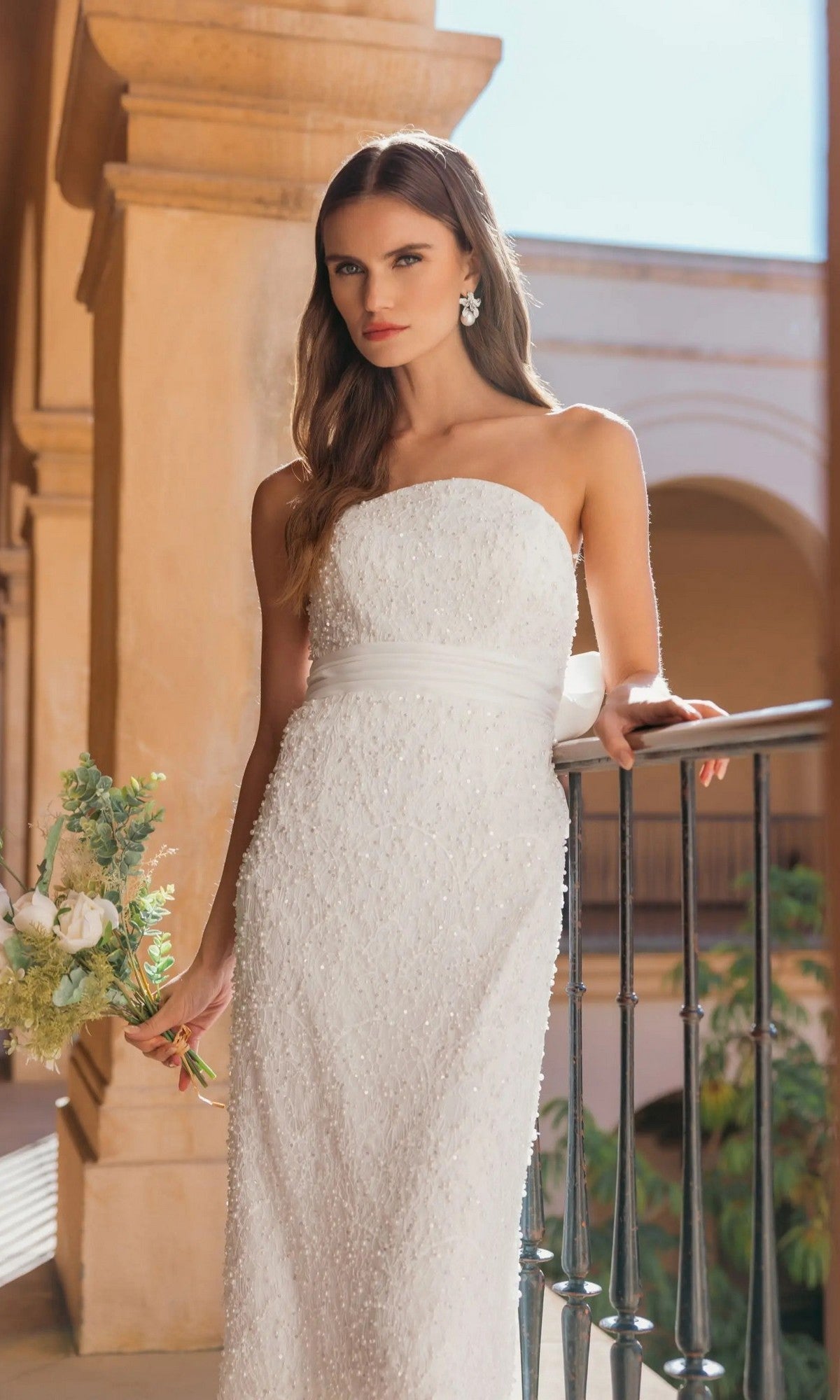 Wearing the Mac Duggal 56416 midi party dress, a woman stands by a railing in an outdoor architectural setting, holding a small bouquet. The strapless white dress features elegant pearl sequin embellishments.