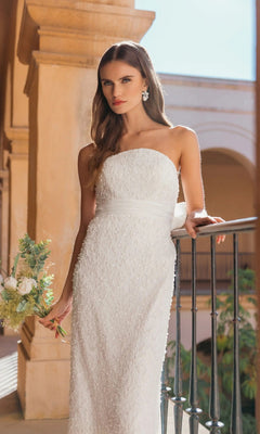 Wearing the Mac Duggal 56416 midi party dress, a woman stands by a railing in an outdoor architectural setting, holding a small bouquet. The strapless white dress features elegant pearl sequin embellishments.