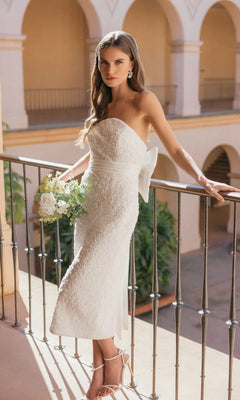 A woman in the Mac Duggal 56416 midi party dress, featuring a strapless silhouette and delicate pearl sequin embellishments, holds a bouquet as she stands on a balcony framed by arches and lush greenery, radiating timeless elegance.