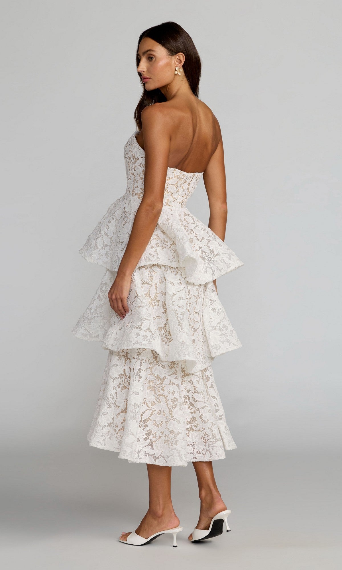 A woman stands sideways against a plain gray background, wearing the Mac Duggal 56419 Tea-Length Party Dress—an ivory strapless lace dress with a ruffled A-line skirt—paired with white heeled mules.