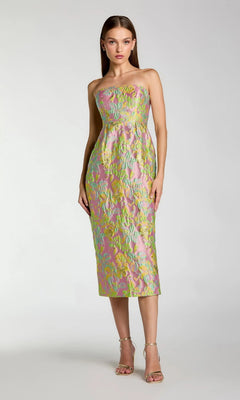 A woman wearing the Midi Wedding Guest Dress: Mac Duggal 56423, a strapless brocade creation in knee-length pastel pink adorned with green and yellow floral patterns, stands against a plain light gray background.