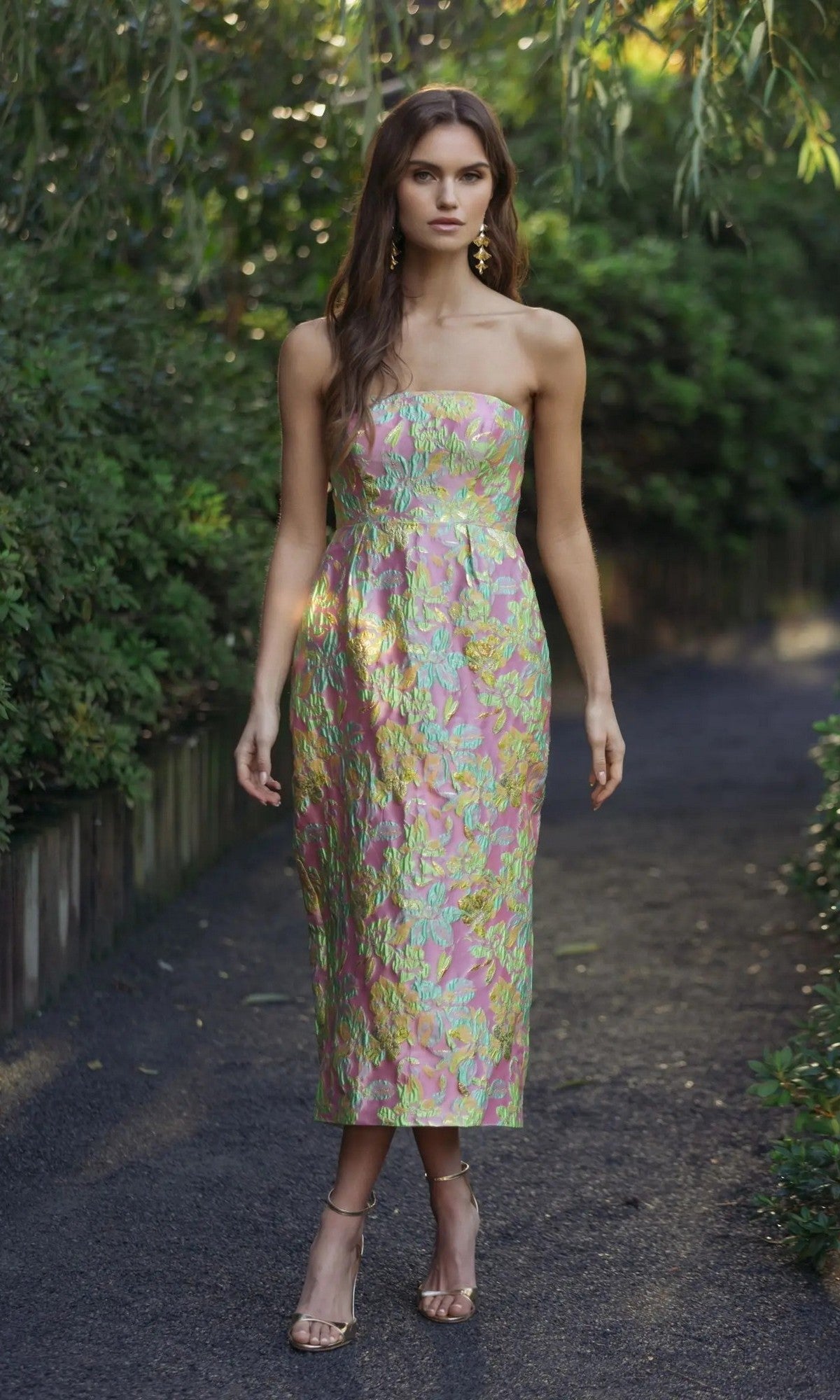 A woman stands on a shaded path wearing the Midi Wedding Guest Dress: Mac Duggal 56423 in Blush Multi—a strapless brocade dress with floral pastel hues—paired with gold heels, with greenery in the background.