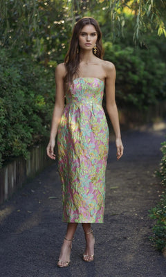 A woman stands on a shaded path wearing the Midi Wedding Guest Dress: Mac Duggal 56423 in Blush Multi—a strapless brocade dress with floral pastel hues—paired with gold heels, with greenery in the background.
