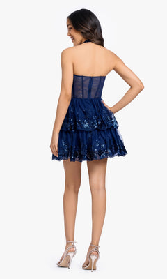 A woman stands facing away, wearing the Periwinkle Short Prom Dress: Blondie Nites 5702BN, a strapless sequin cocktail dress with tiered layers, paired with silver high-heeled sandals.