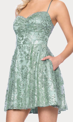 A woman confidently poses with one hand in the pocket of the Mini Green Sequin Hoco Dress: Blondie Nites 5705BN, featuring thin straps and a flared skirt—perfect for junior sizes.