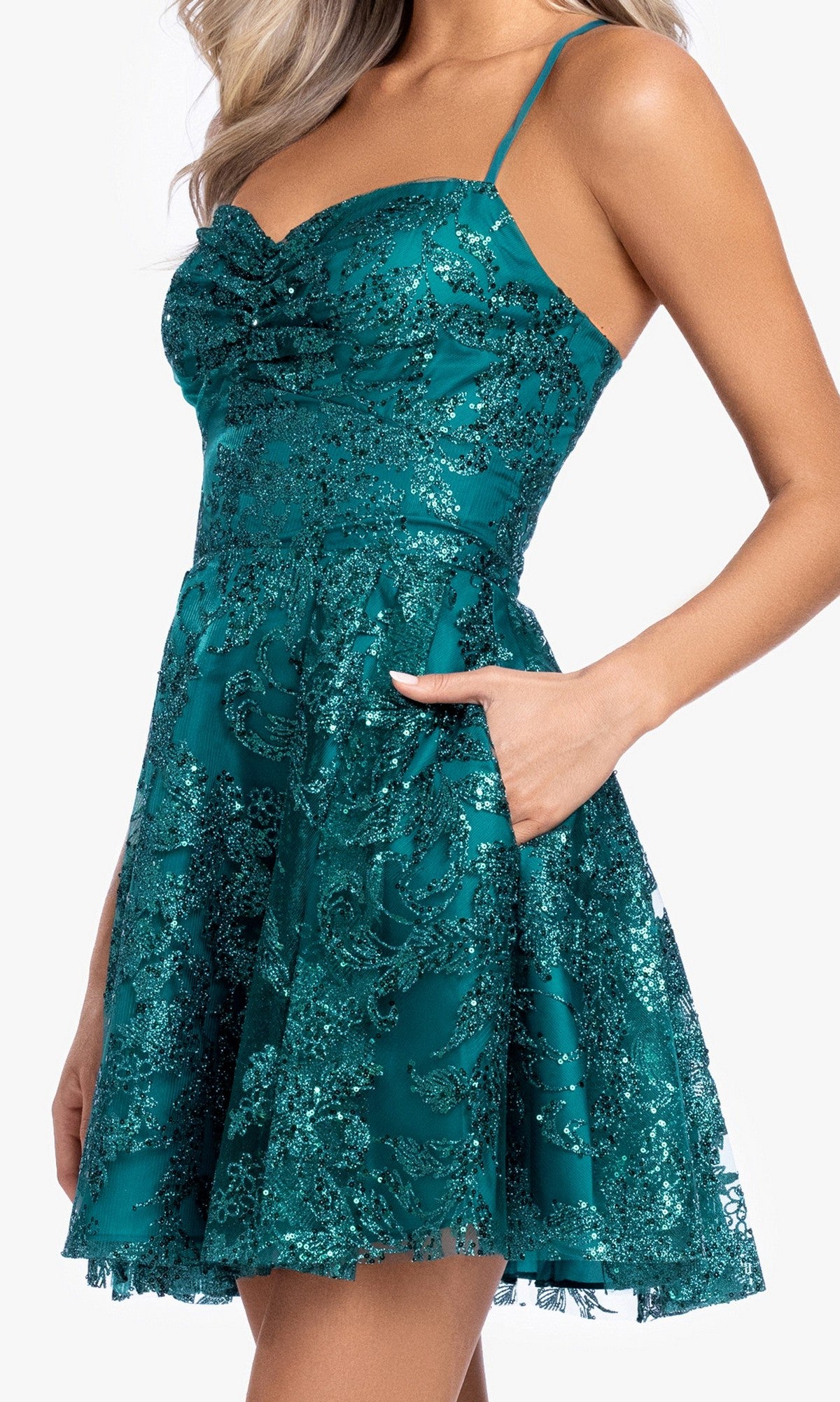 A woman stands with one hand in the pocket, wearing the Mini Green Sequin Hoco Dress: Blondie Nites 5705BN, featuring thin straps, a fitted bodice, flared skirt, and glittery floral patterns.