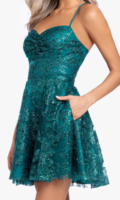 A woman stands with one hand in the pocket, wearing the Mini Green Sequin Hoco Dress: Blondie Nites 5705BN, featuring thin straps, a fitted bodice, flared skirt, and glittery floral patterns.