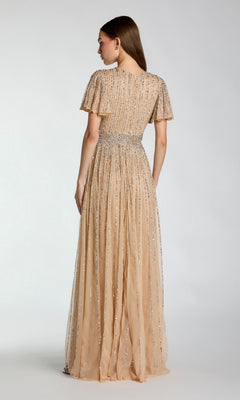 A woman is seen from the back, standing against a plain light background, wearing the Mac Duggal 5713 Long Formal Dress—an elegant peach, floor-length gown with short sleeves and vertical silver embellishments.