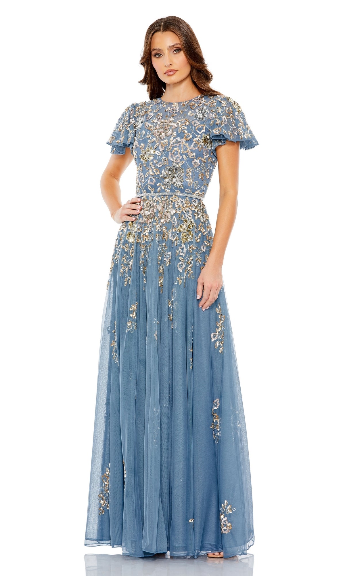 A woman models the Mac Duggal 5717 Short Sleeve Beaded Formal Dress, a floor-length blue A-line gown with gold floral embroidery and flutter sleeves, standing against a plain white background.
