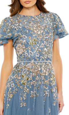 A woman is seen from the head to upper body wearing the Mac Duggal 5717, a short sleeve beaded formal dress in blue with sheer fabric and elaborate gold, white, and silver floral embroidery.
