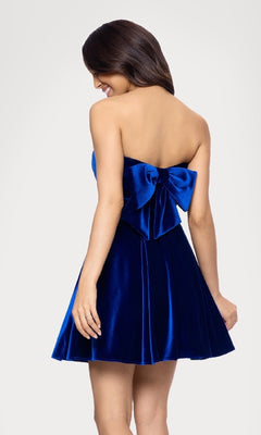 A woman stands against a plain light background wearing the Short Velvet Hoco Dress: Blondie Nites 5718BN, a blue strapless homecoming dress with a large back bow.