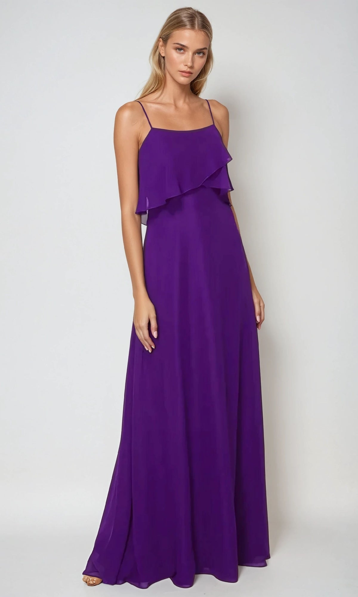 A woman models the Juliet 591, a sleeveless, floor-length purple wedding-guest dress with spaghetti straps and a ruffled pop-over bodice, standing against a plain light background.