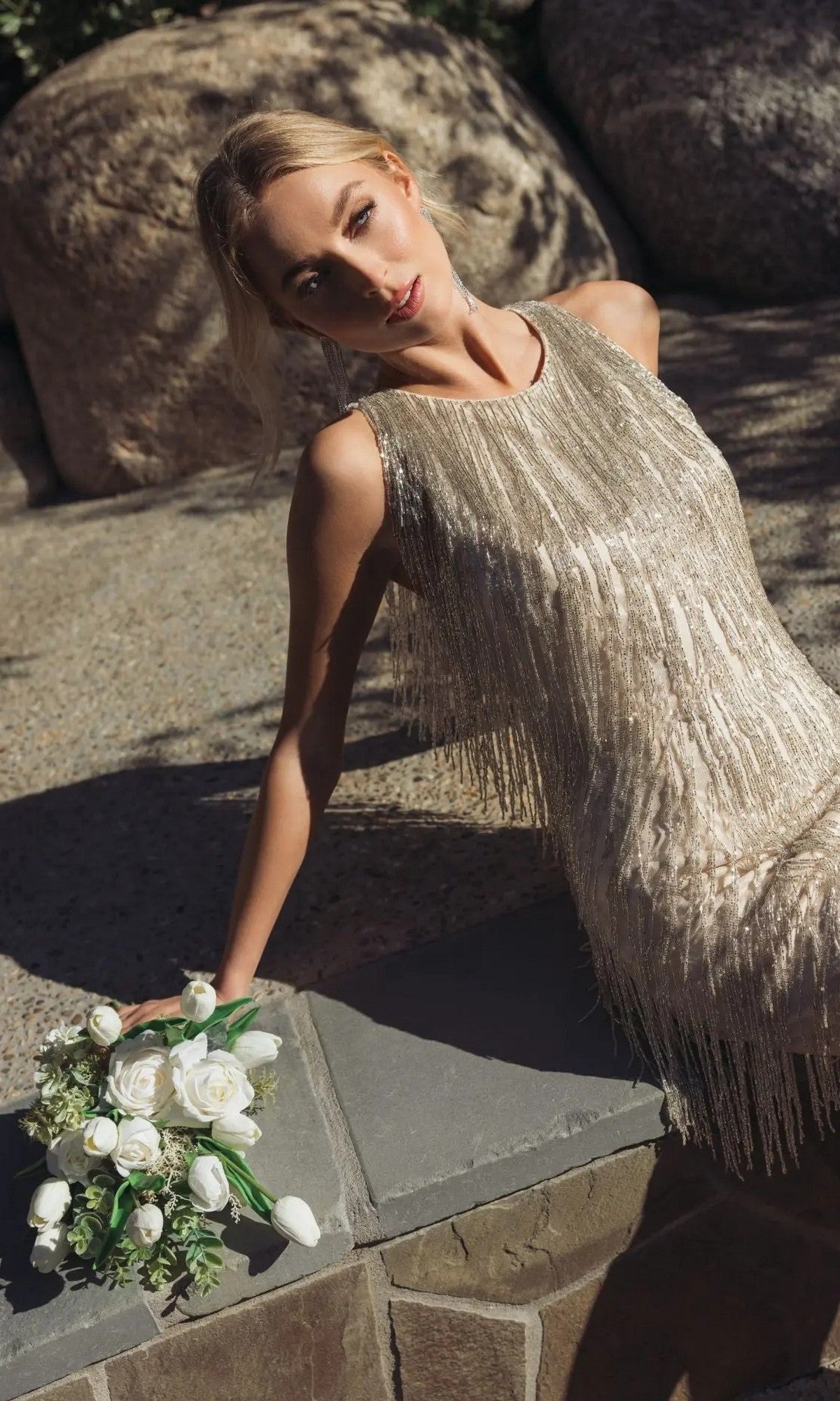 A woman in the Short Beaded Fringe Party Dress: Mac Duggal 5934 leans against a stone surface outdoors, holding a small bouquet of white roses.