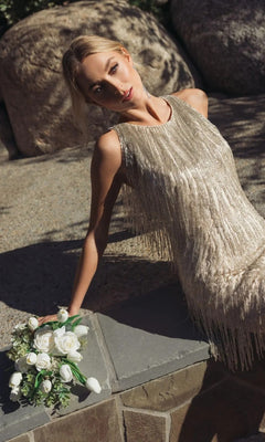 A woman in the Short Beaded Fringe Party Dress: Mac Duggal 5934 leans against a stone surface outdoors, holding a small bouquet of white roses.