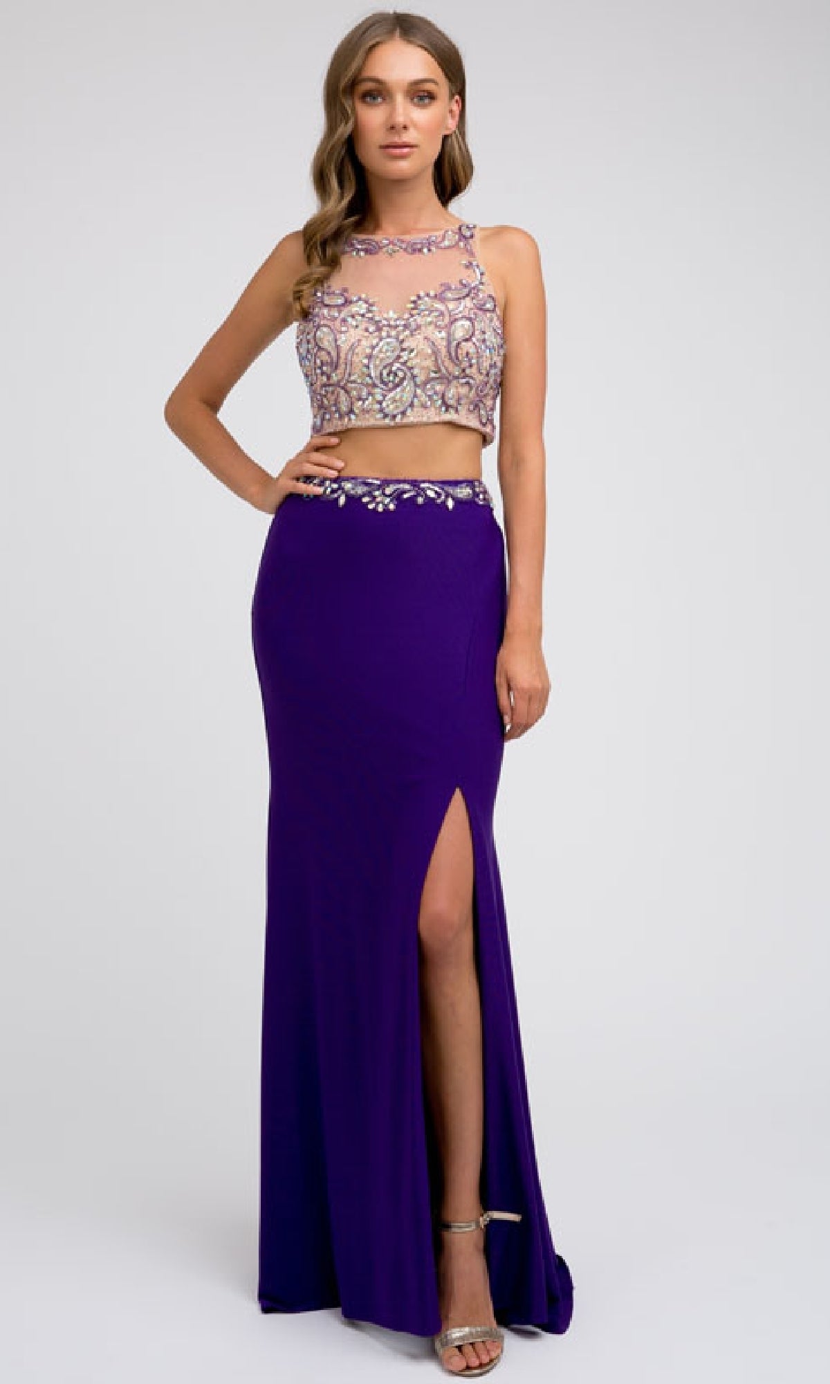 A woman wears the Juliet 595 Long Prom Dress—a sleeveless, beaded top paired with a purple skirt featuring a side leg slit and silver sandals—for an elegant two-piece prom look against a plain background.
