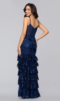 A woman in the Blondie Nites 5962BN long prom dress, featuring navy blue sequins and a tiered ruffle skirt, stands slightly turned to the side and smiling against a plain light gray background.