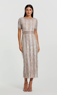 A woman models the Mac Duggal 5980 Midi Fringe Wedding-Guest Dress, featuring short sleeves, silver sequins, and fringe, paired with silver high-heeled sandals—an eye-catching semi-formal style against a plain backdrop.