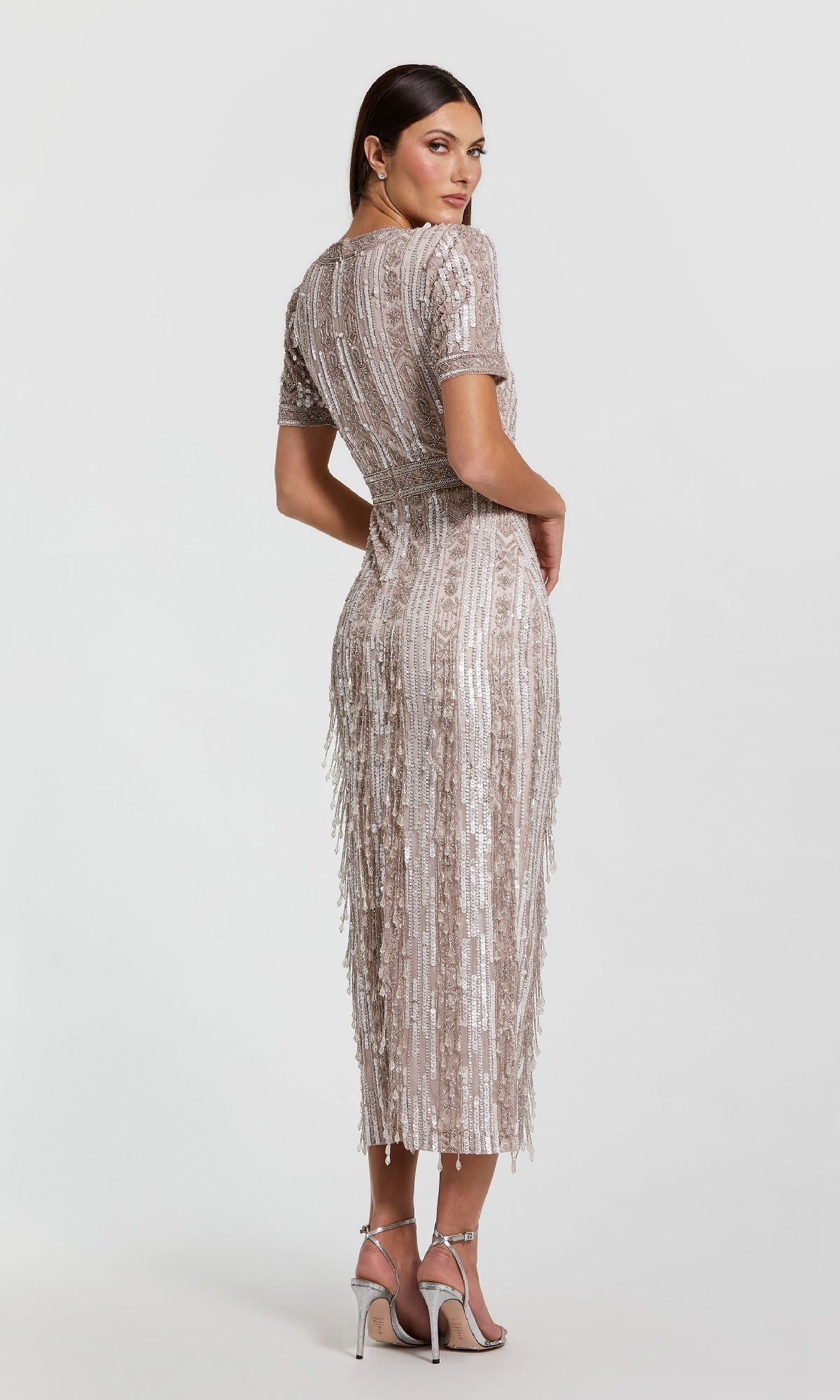Wearing the Mac Duggal 5980 Midi Fringe Wedding-Guest Dress, a woman stands in profile, looking over her shoulder. The beaded, short sleeve dress with fringe details is paired with silver high-heeled sandals.