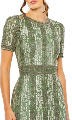 A woman stands with one arm at her side, wearing the Mac Duggal 5980 Midi Fringe Wedding-Guest Dress in green, short-sleeved and adorned with intricate white beadwork and geometric patterns.