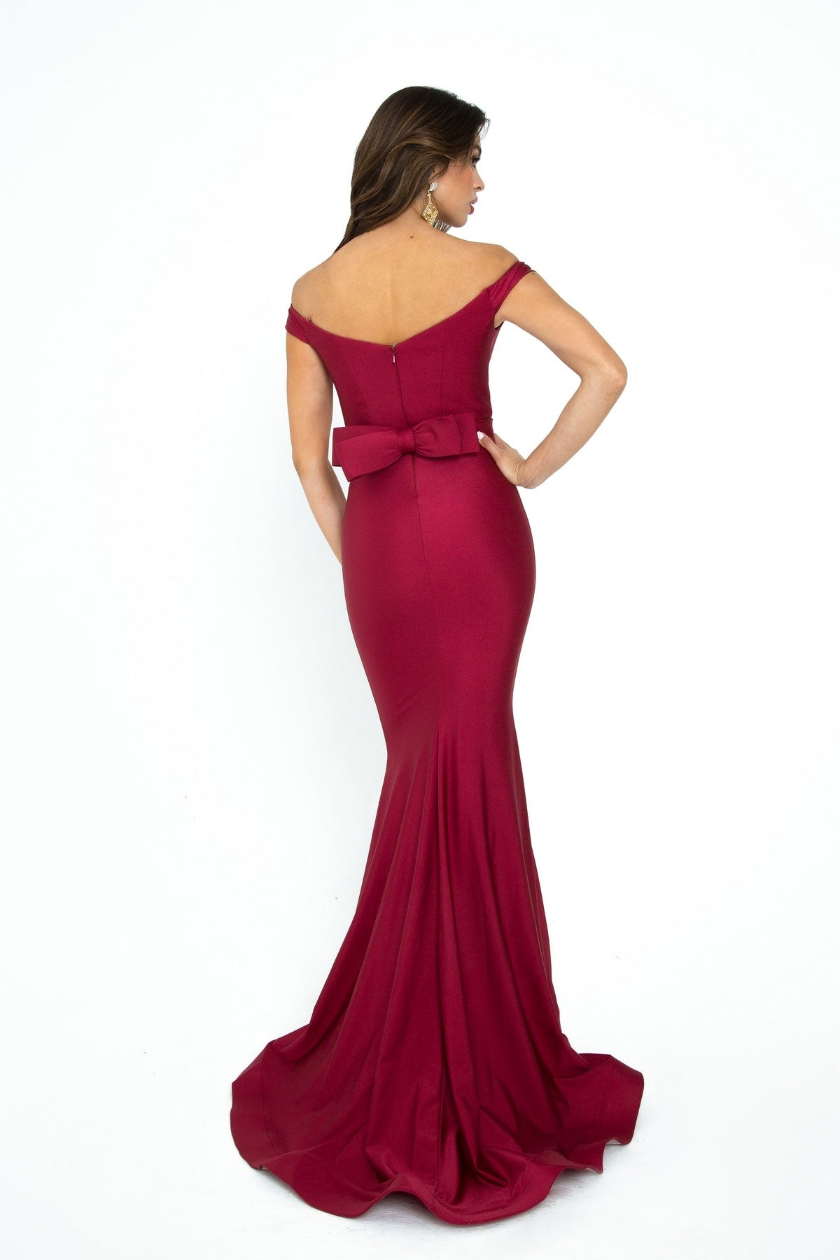 Woman wearing the Atria Off-the-Shoulder Long Formal Dress 6016H in burgundy with waist bow detailing, standing against a plain white background—an elegant option for prom or evening occasions.